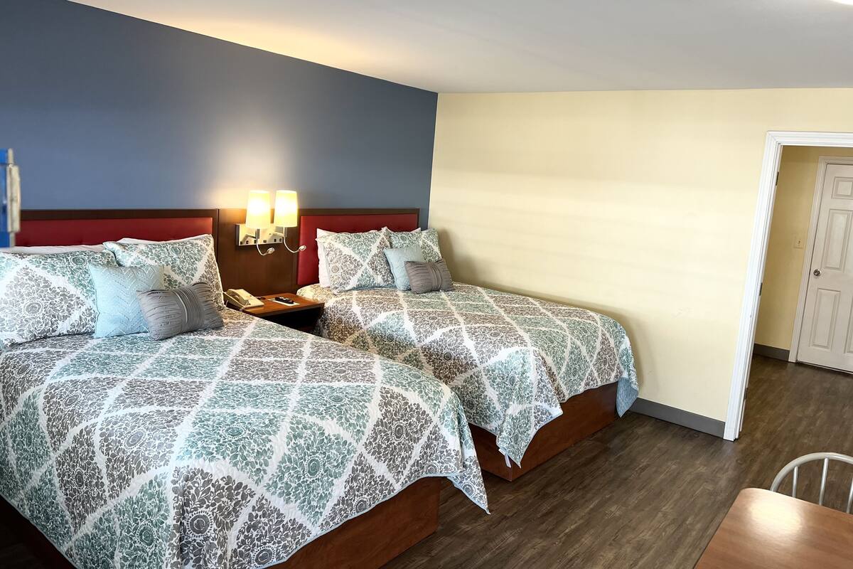 Habitación de hotel con dos camas, una mesita de noche con lámpara y una puerta que da a otra habitación.