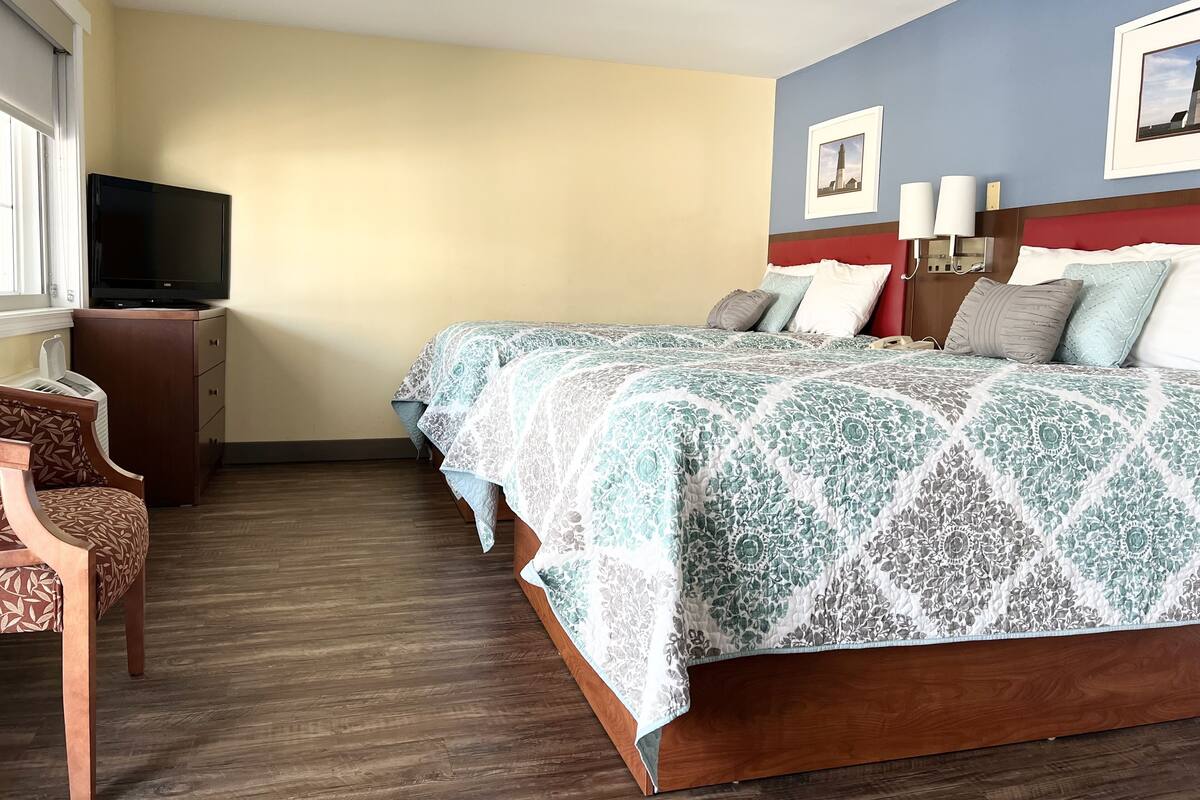 Una habitación de hotel con una cama grande, televisión, mesita de noche de madera y una silla.