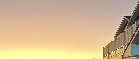 A coastal building with a balcony overlooking the beach, a parked car, and a sunset in the background.