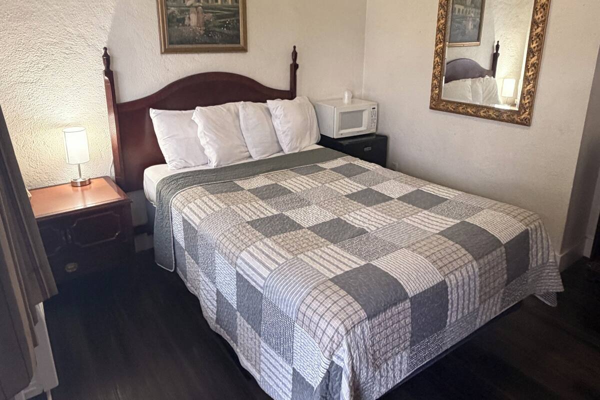 A neatly made bed with a patterned quilt, a bedside lamp, a framed painting, a bedside table with a microwave, and a mirror on the wall.