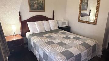 A neatly made bed with a patterned quilt, a bedside lamp, a framed painting, a bedside table with a microwave, and a mirror on the wall.