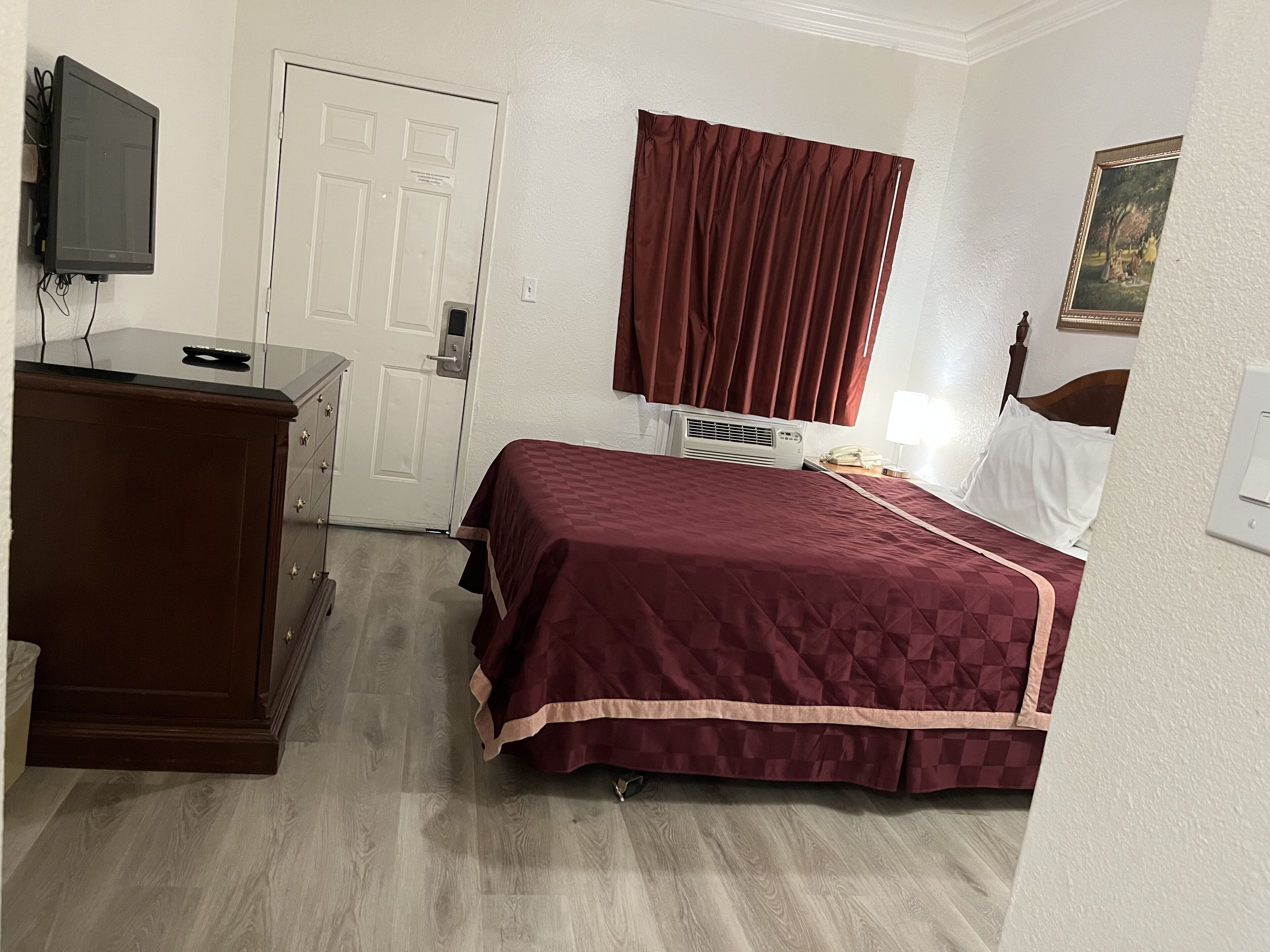 Quarto de hotel com cama, penteadeira, televisão, janela com cortinas e um quadro na parede.