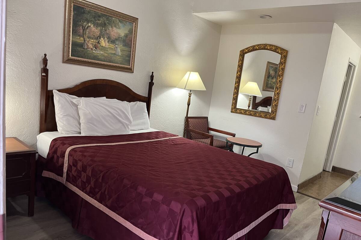 A hotel room with a large bed, a nightstand, a chair, a small table, a lamp, a painting, and a mirror.
