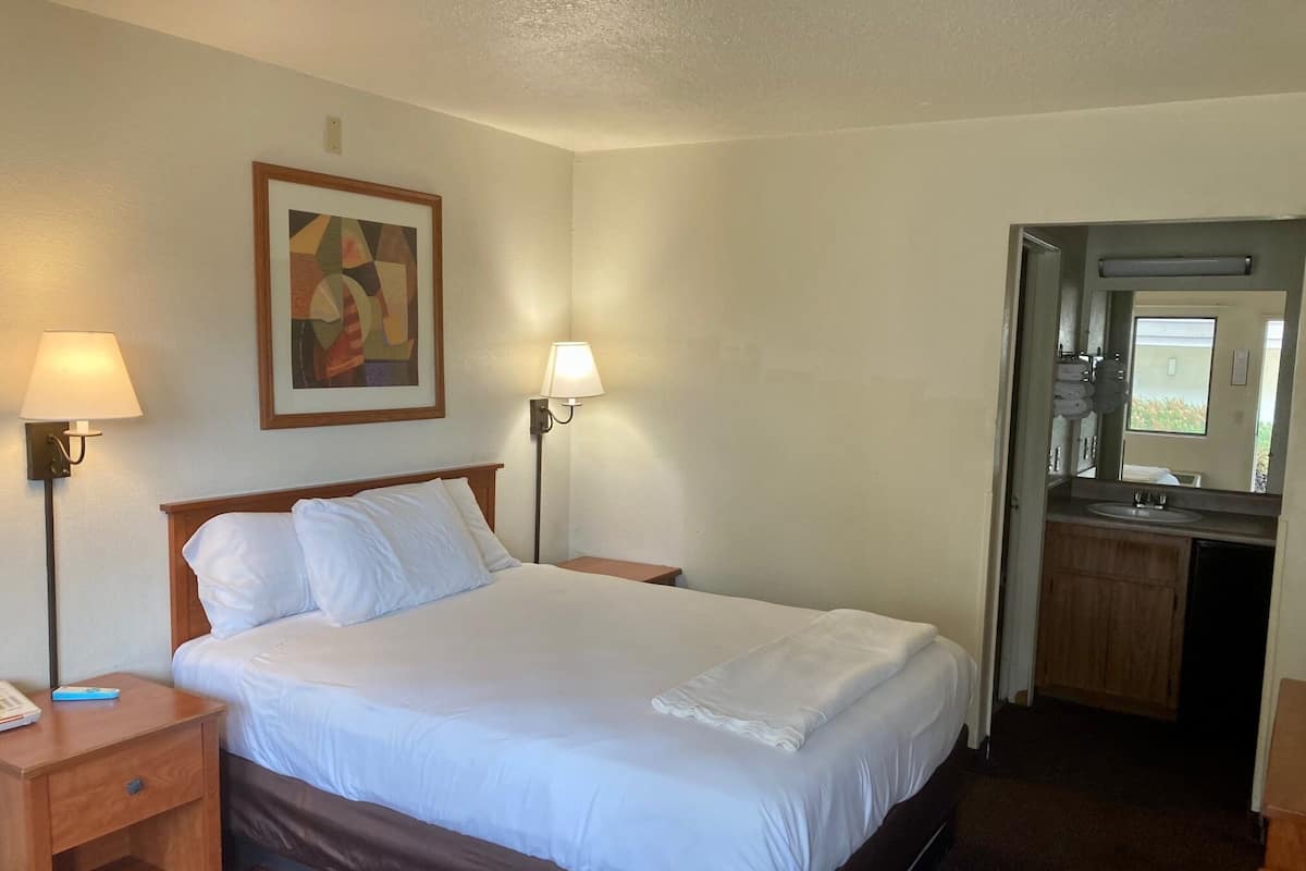 A hotel room with a bed, two bedside lamps, a painting on the wall, a wooden nightstand, and a bathroom visible through an open door.