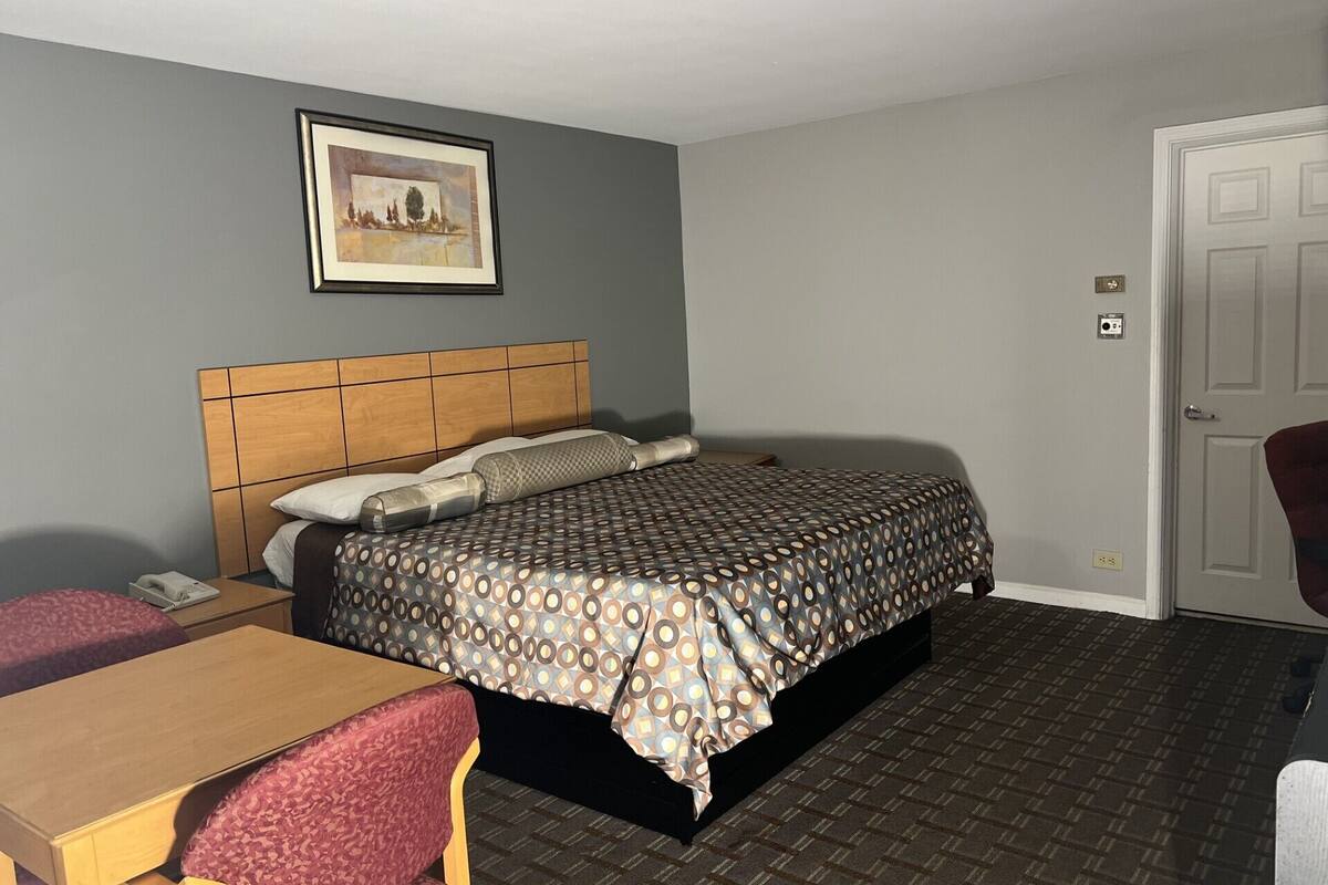 A hotel room with a bed, a wooden headboard, a desk with a chair, a framed picture on the wall, and a door.