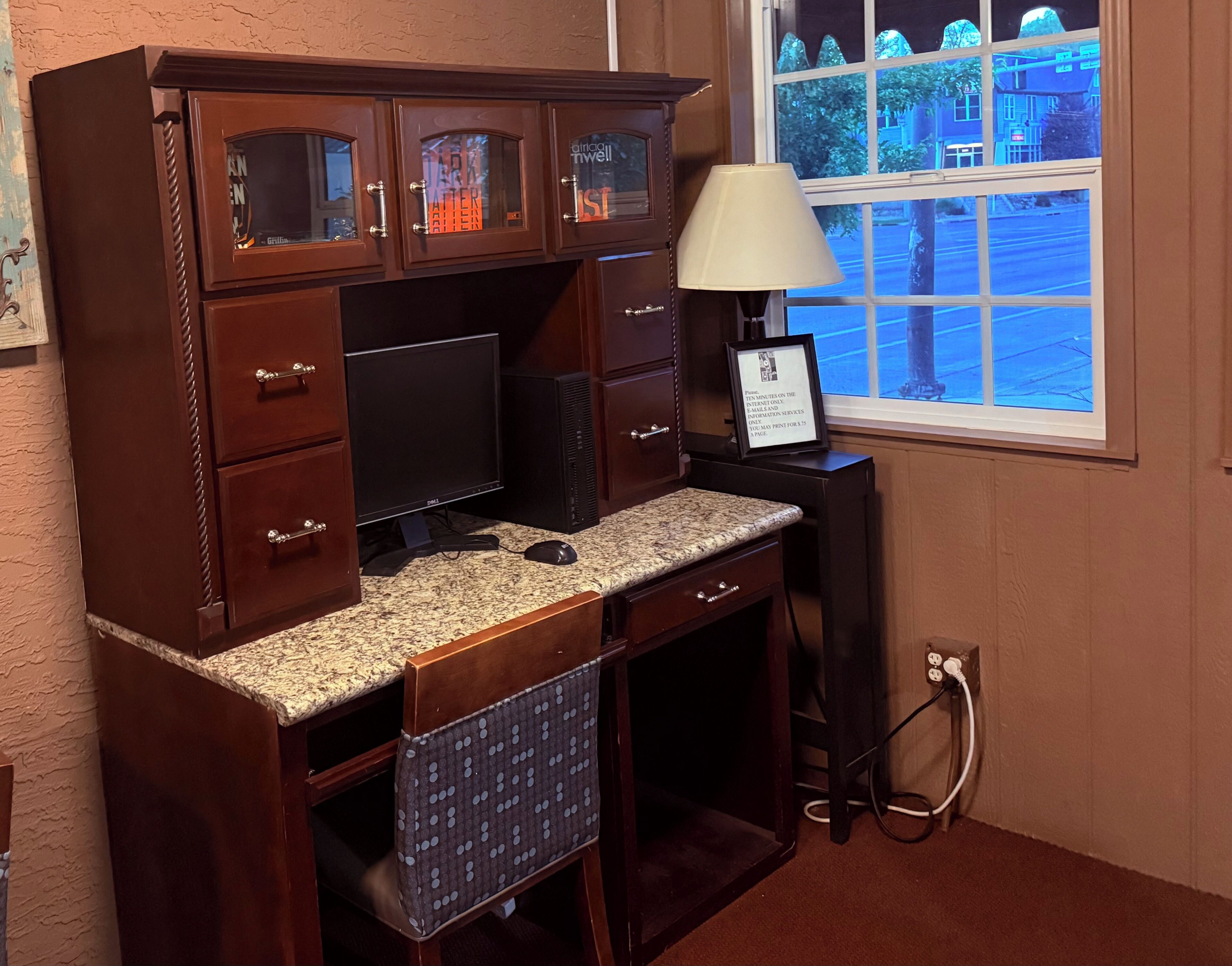 A wooden desk with a computer, a lamp, and a window with a view of trees and buildings.
