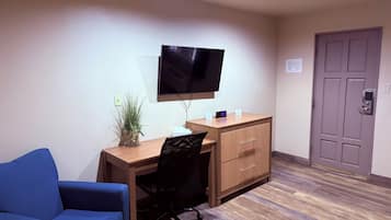 Habitación de hotel con un escritorio de madera, una silla de oficina negra, un televisor, una planta en maceta, un sofá azul y una puerta con cerradura.