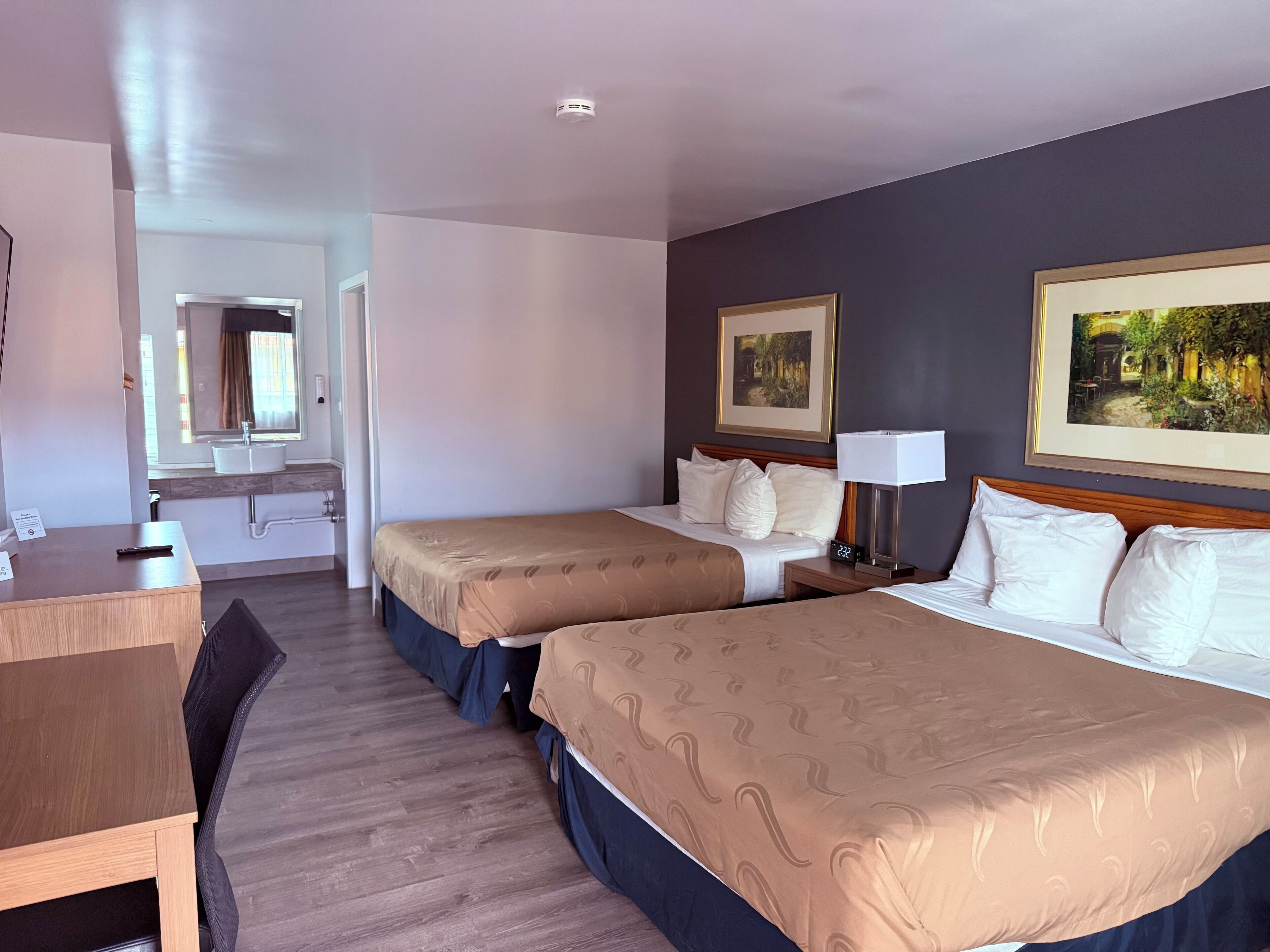 Una habitación de hotel con dos camas, un escritorio, una silla, una mesita de noche con una lámpara, un espejo y dos cuadros en la pared.