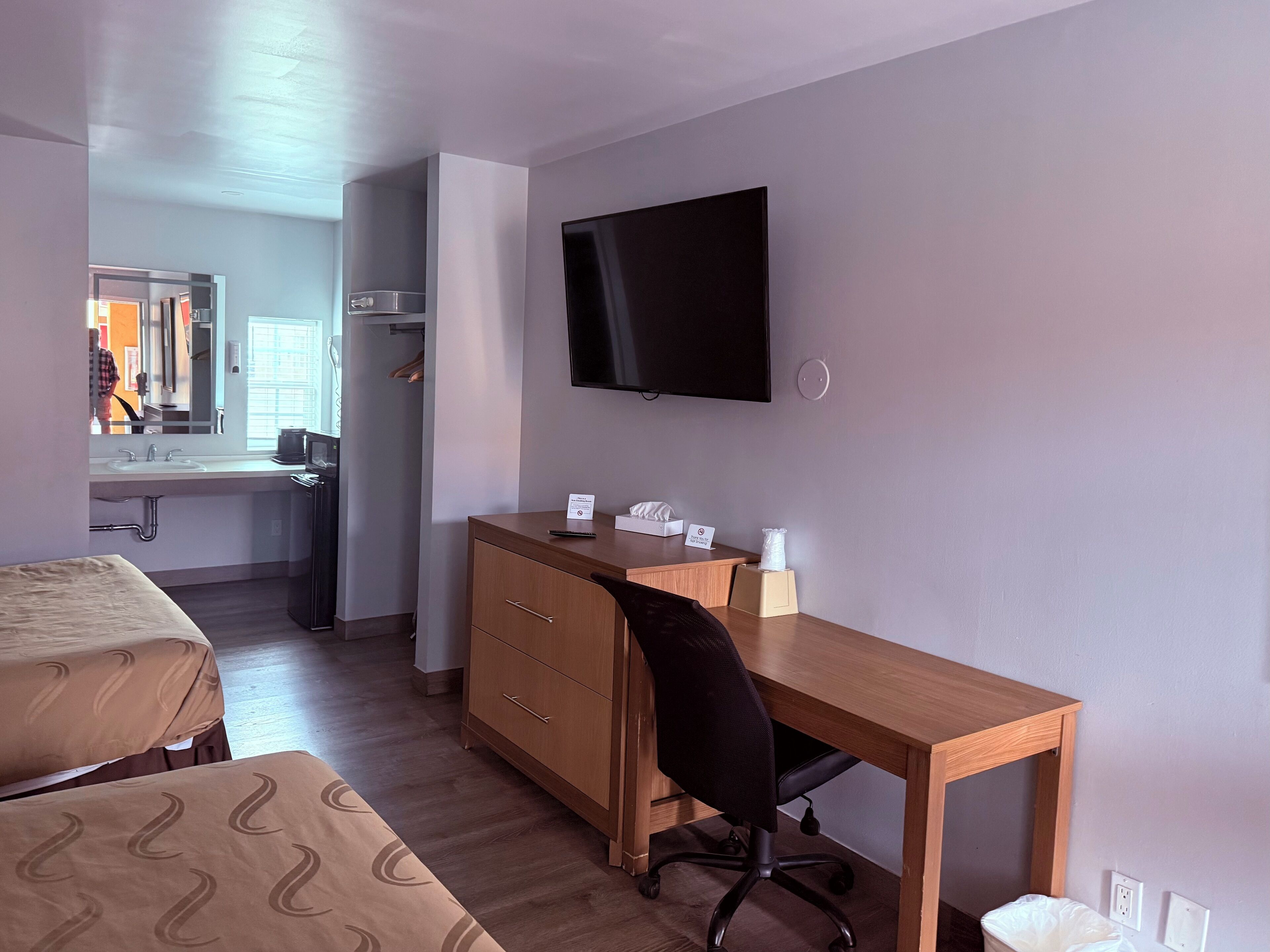 A hotel room with a bed, a desk, a chair, a TV, a mirror, and a bathroom.