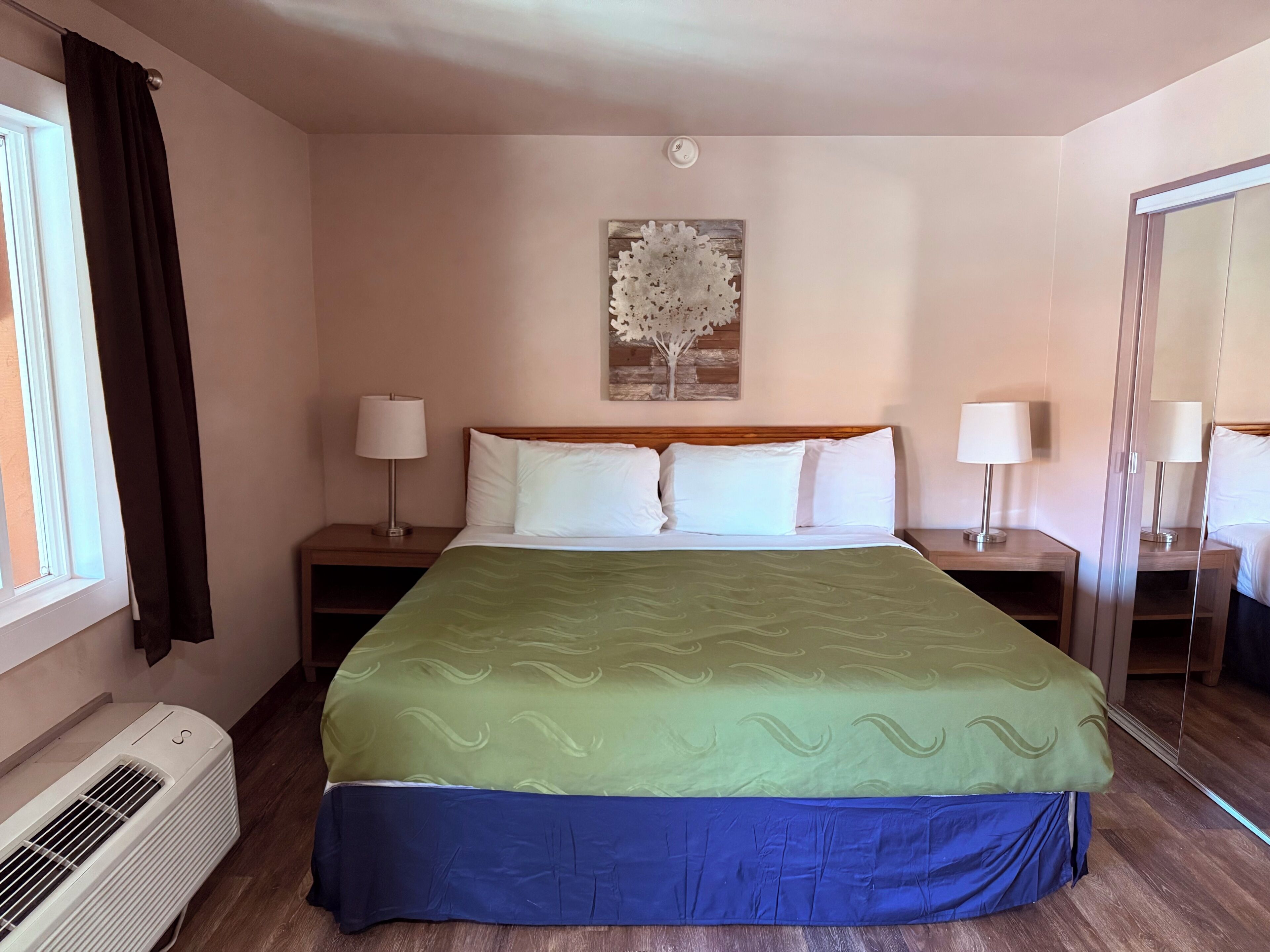 A hotel room with a bed, two bedside tables with lamps, a wall-mounted air conditioner, and a window with curtains.