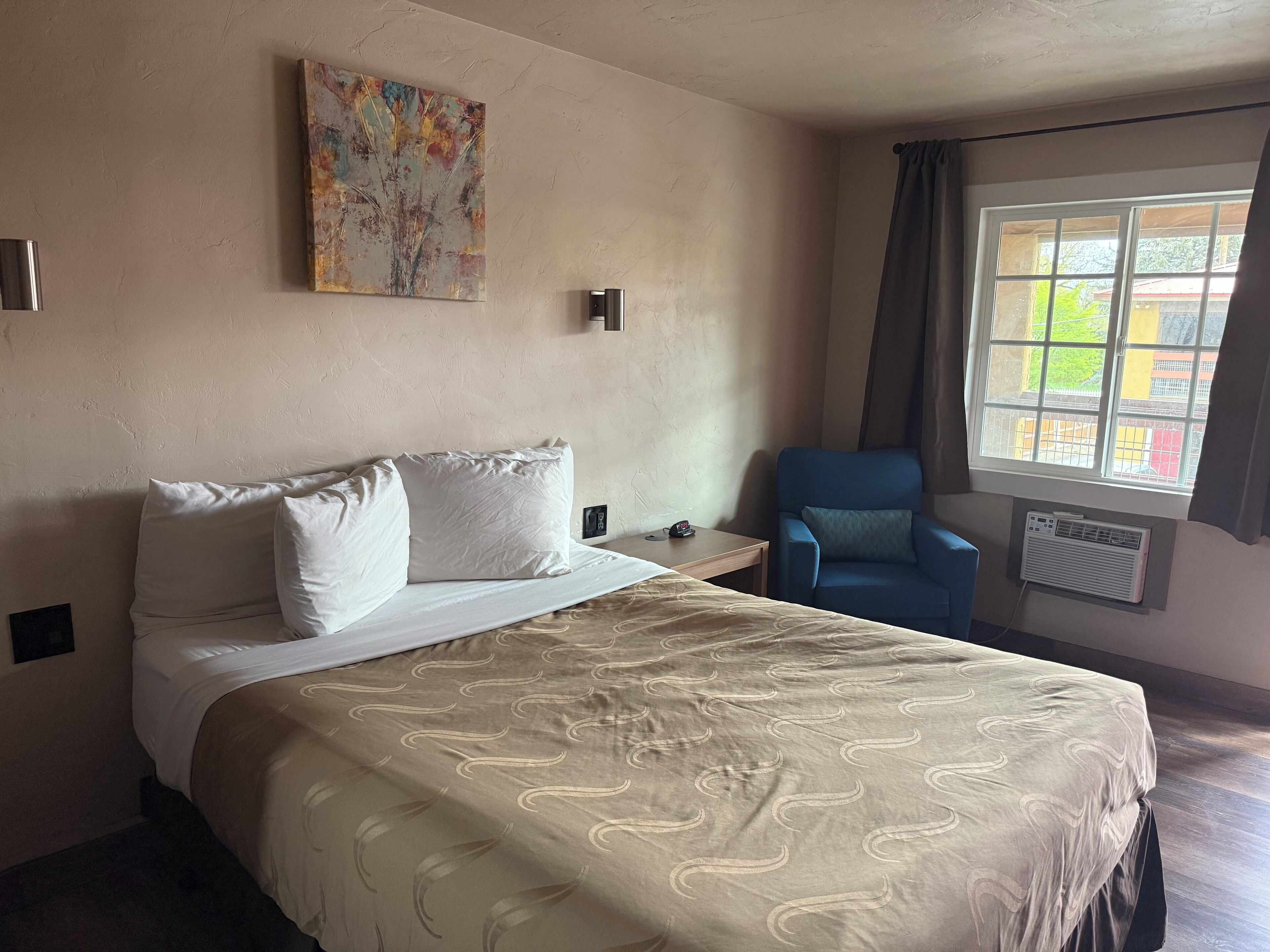 A hotel room with a bed, a chair, a window with curtains, and a wall-mounted air conditioner.