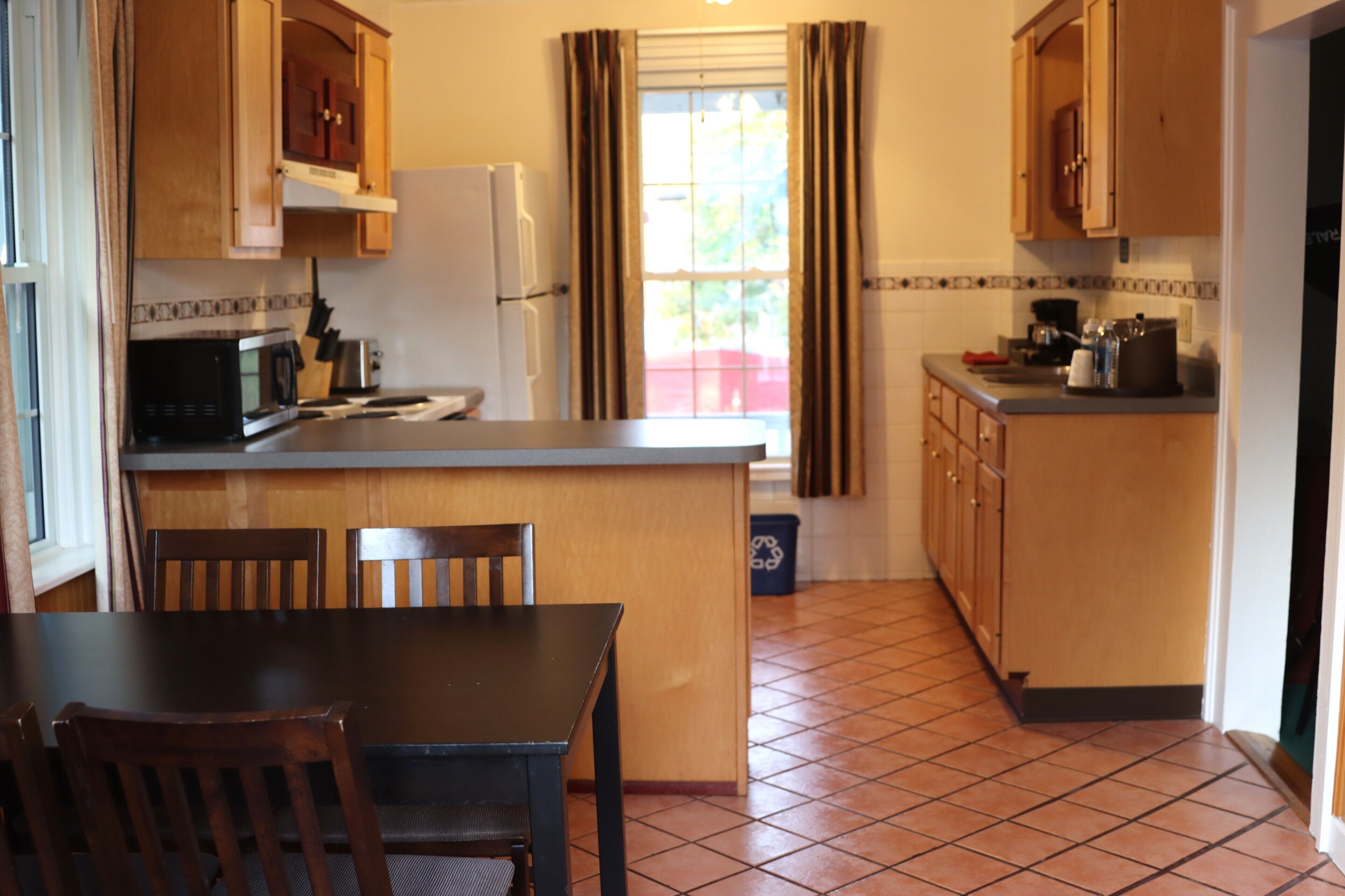 A kitchen with a dining table, chairs, a refrigerator, and a window with curtains.