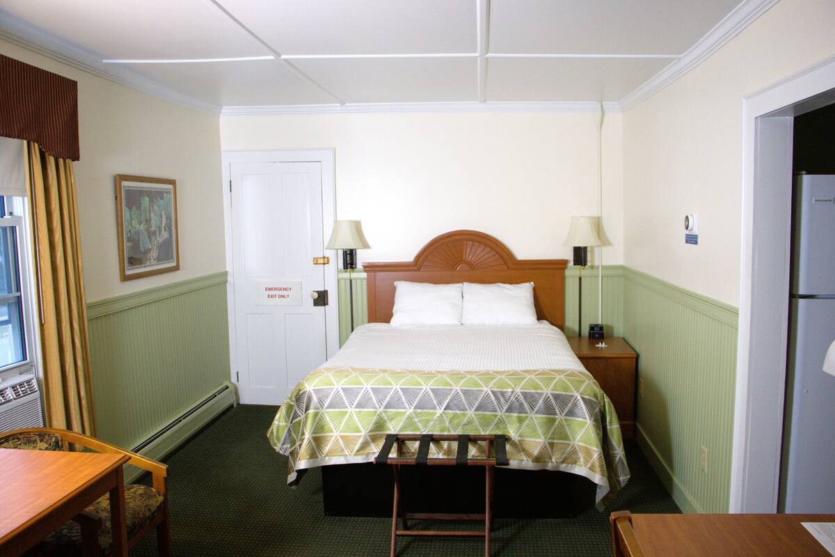 Une chambre d’hôtel comprenant un lit, des tables de chevet, une chaise, une fenêtre avec des rideaux et une porte portant l’inscription « Sortie de secours uniquement ».