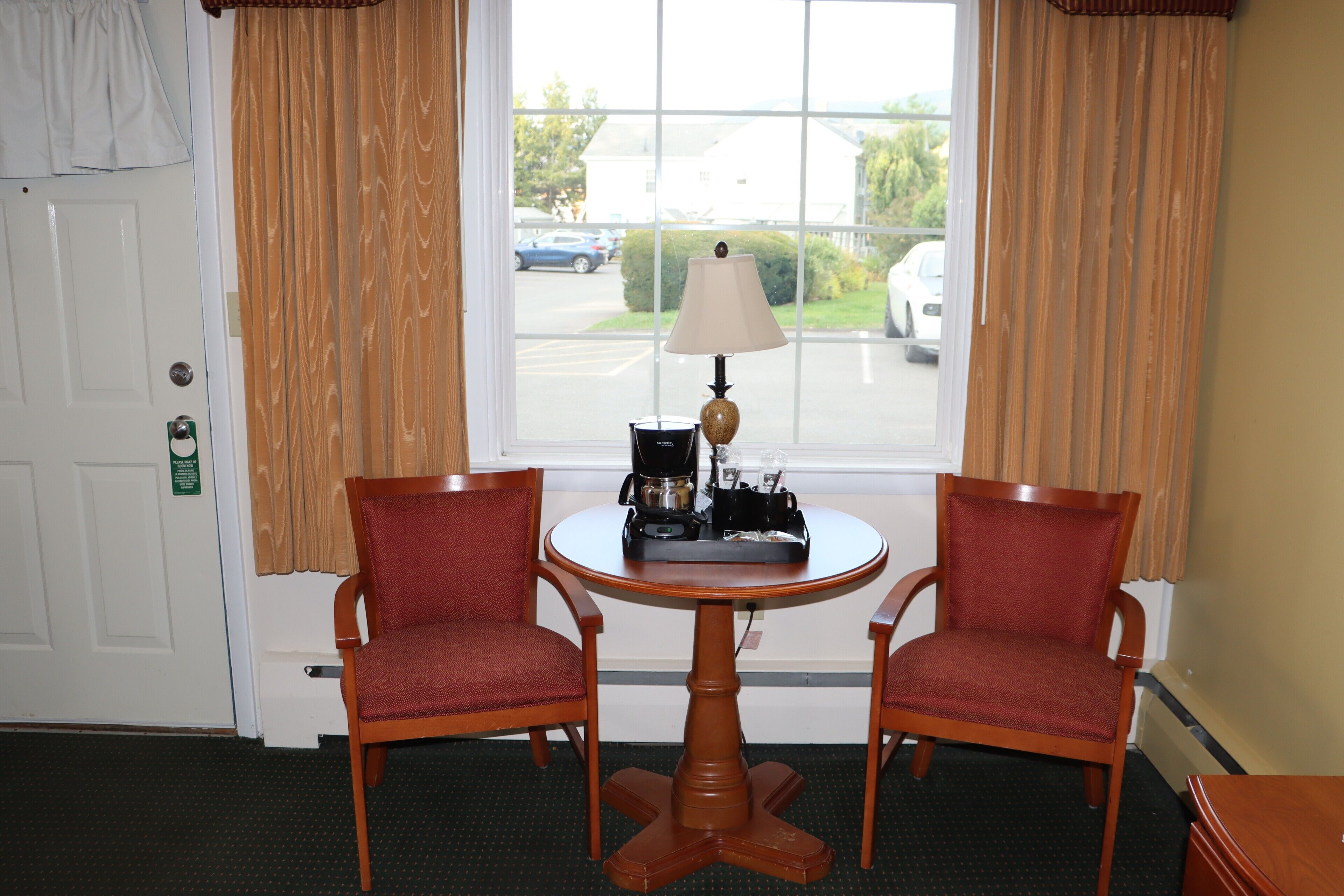 A room with two wooden chairs around a small round table with a lamp, a window with curtains, and a door with a sign.