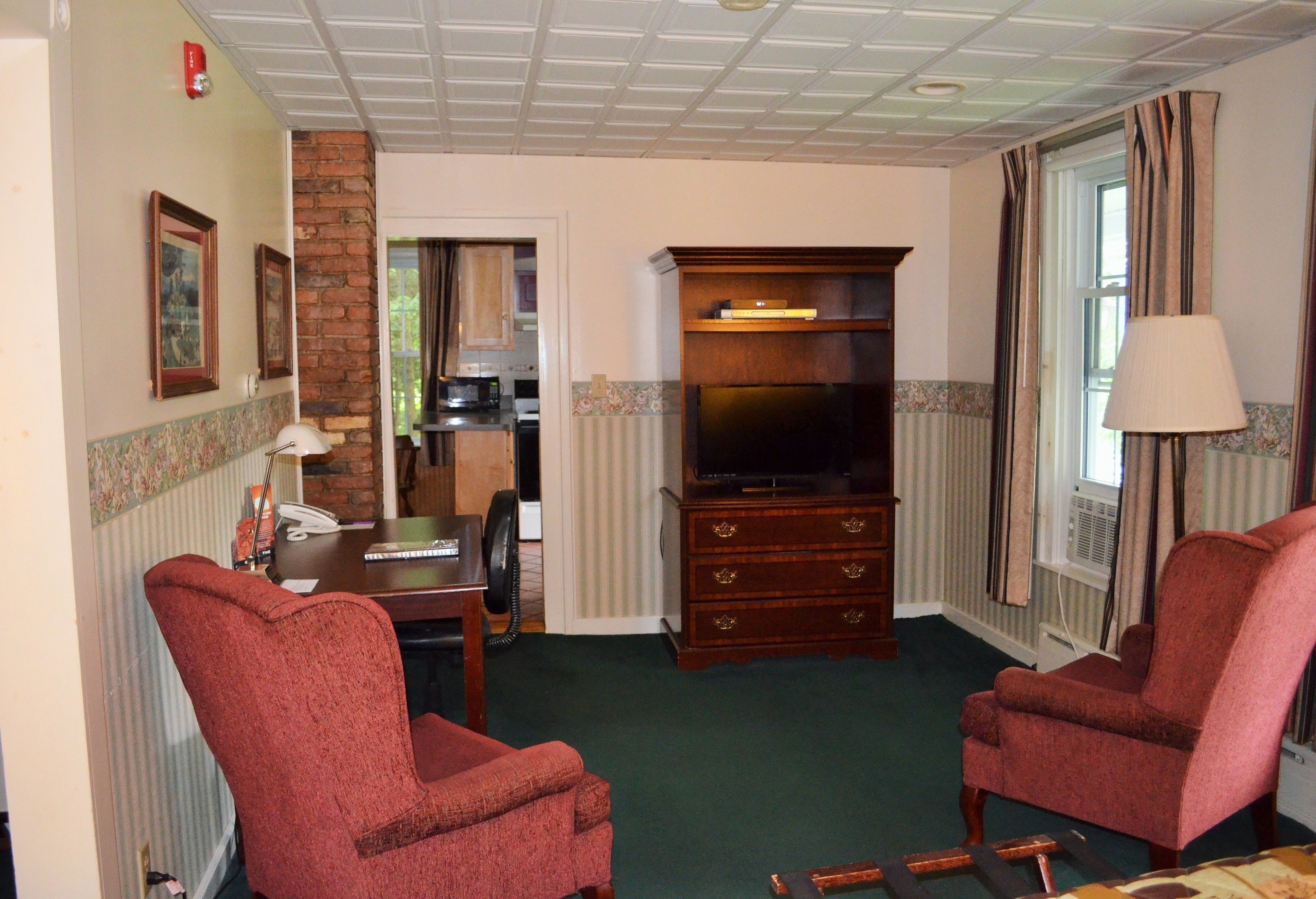 A living room with two red armchairs, a wooden cabinet with a television, a desk with a telephone, and a lamp.