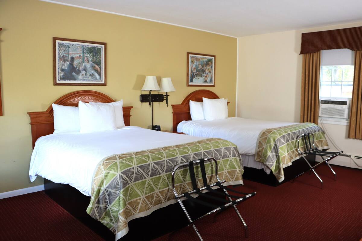 Une chambre d’hôtel avec deux lits, une tête de lit en bois, une moquette rouge, deux lampes fixées au mur et deux tableaux accrochés au mur.