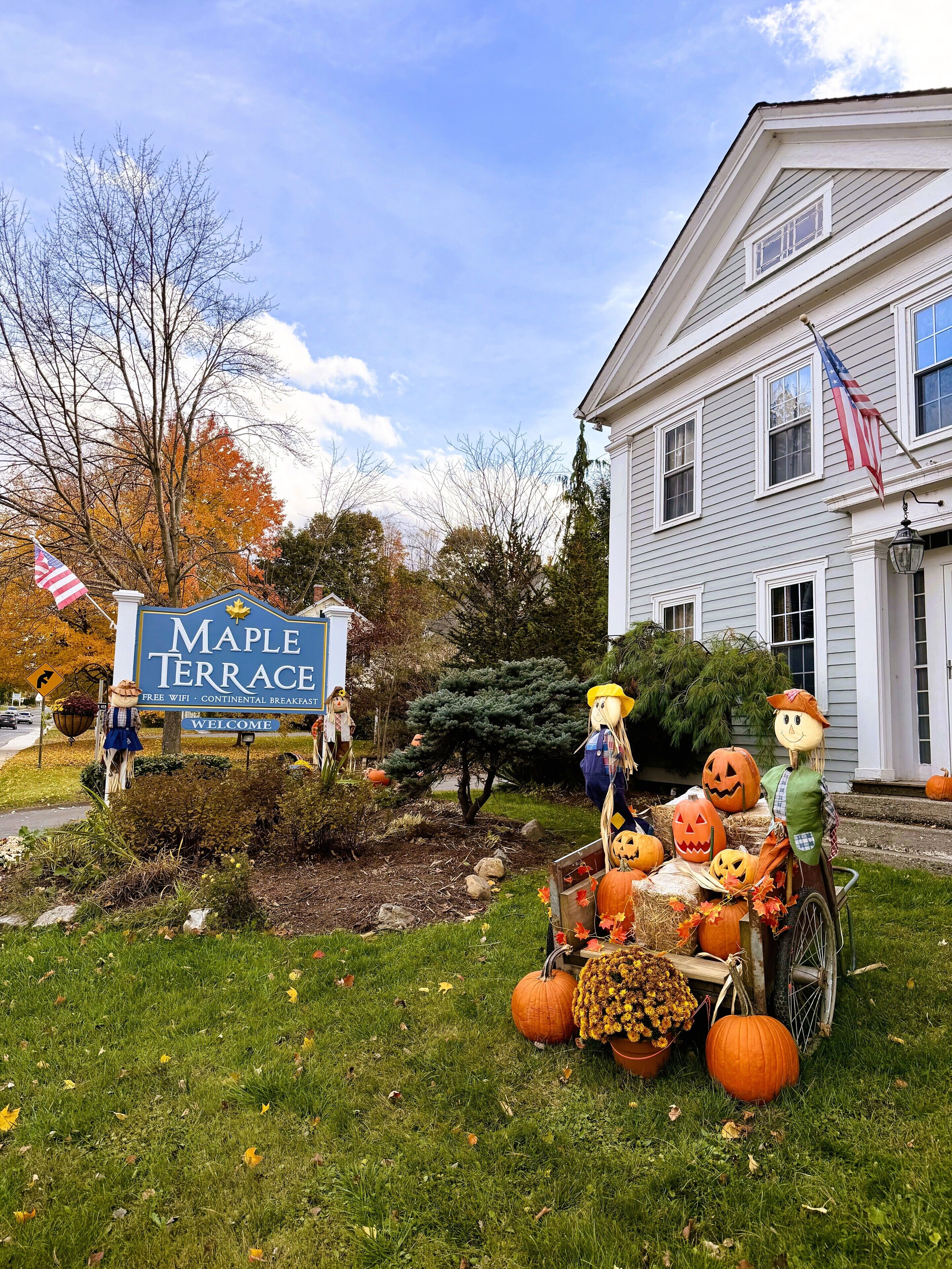 A sign for Maple Terrace with a welcome message, a lawn with autumn leaves, and a Halloween decoration with pumpkins and scarecrows.