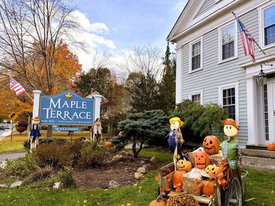 A sign for Maple Terrace with a welcome message, a lawn with autumn leaves, and a Halloween decoration with pumpkins and scarecrows.