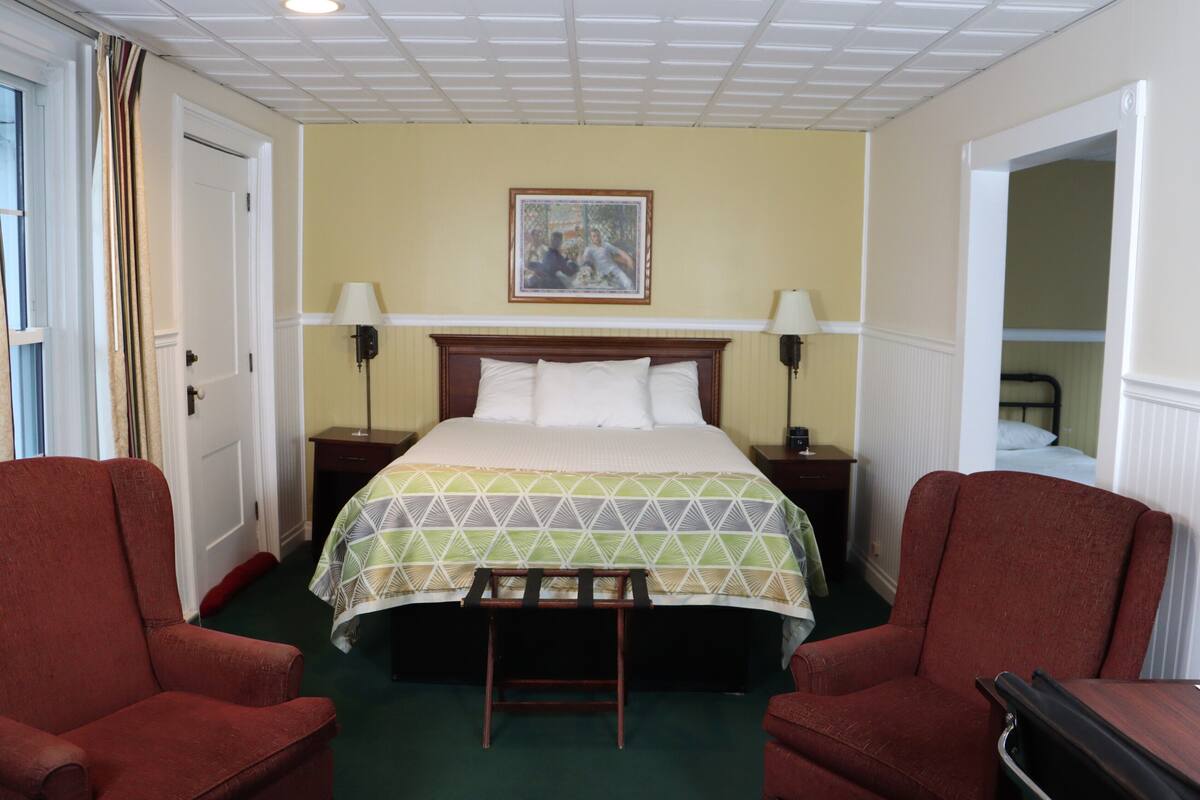 Une chambre d’hôtel comprenant un lit, deux tables de chevet, deux lampes, un banc et deux fauteuils.