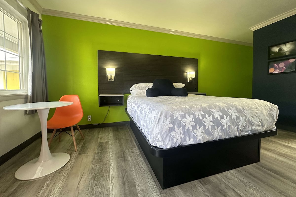 A hotel room with a bed, a small table, an orange chair, and a green accent wall.