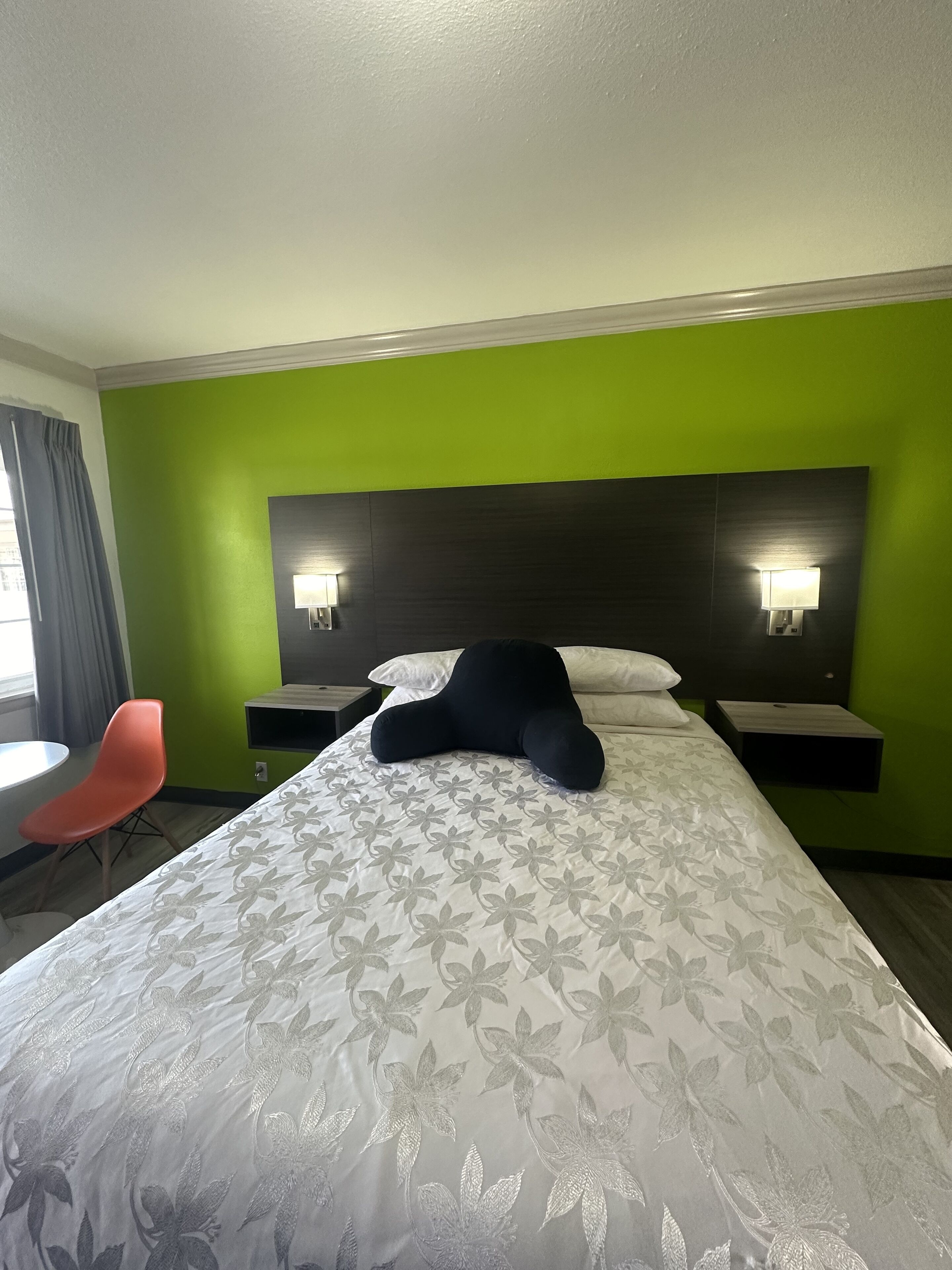 A hotel room with a green wall, a large bed with a floral-patterned bedspread, two bedside tables with lamps, a red chair, and a window with curtains.