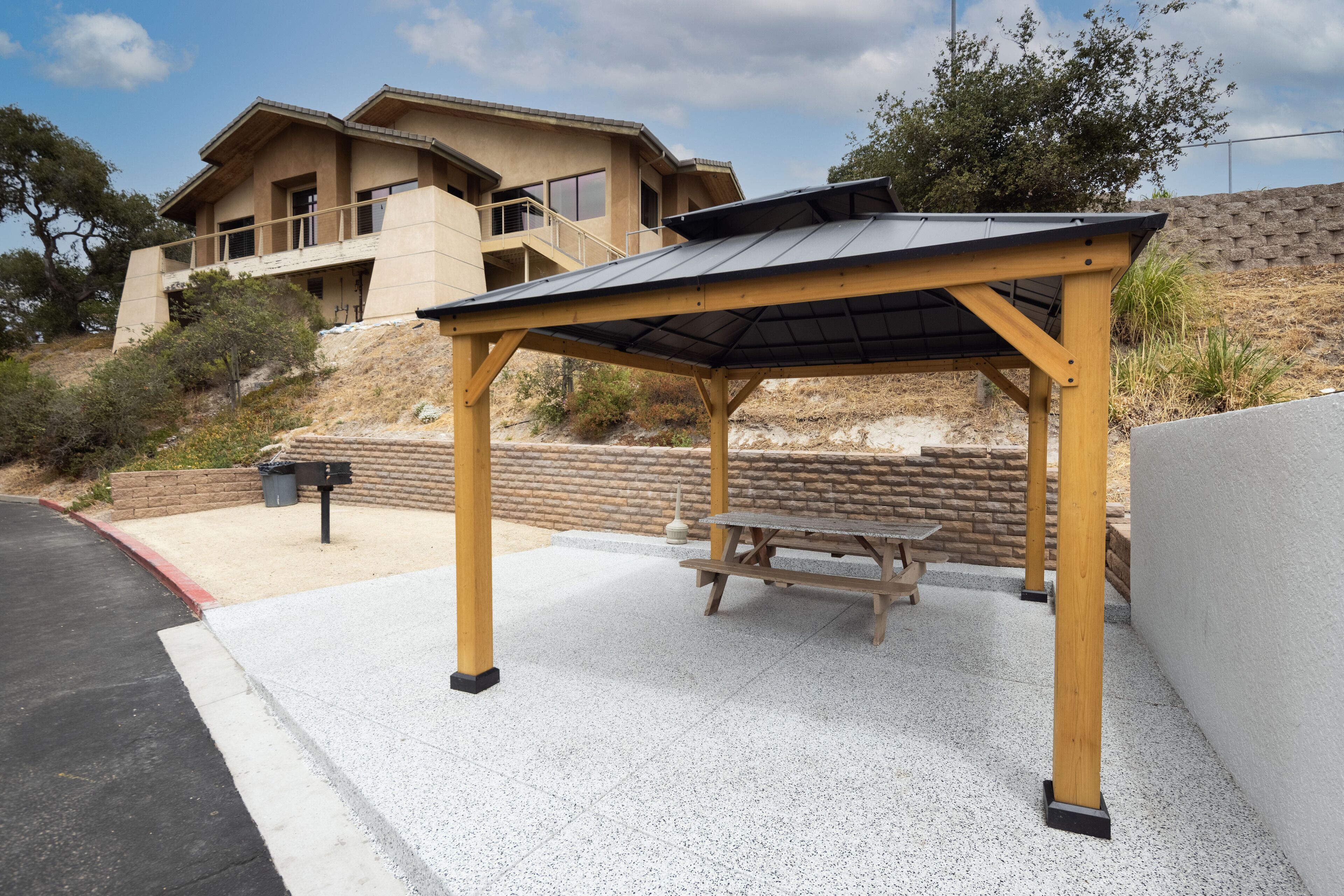 A covered outdoor seating area with a picnic table, adjacent to a building with a balcony.