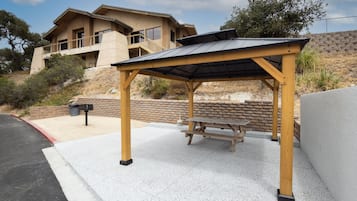 A covered outdoor seating area with a picnic table, adjacent to a building with a balcony.