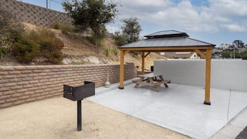 Een overdekt terras met een picknicktafel en banken, een brievenbus, en een bakstenen muur op de achtergrond.