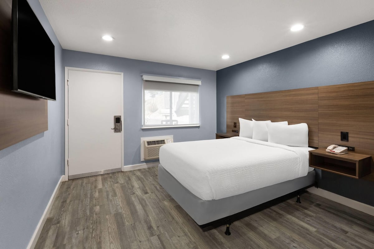 A hotel room with a large bed, a bedside table with a phone, a wall-mounted TV, and a window with blinds.