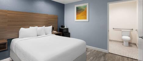 Un dormitorio moderno con una cama grande, cabecera de madera, un cuadro enmarcado en la pared, y un baño con inodoro y lavabo.