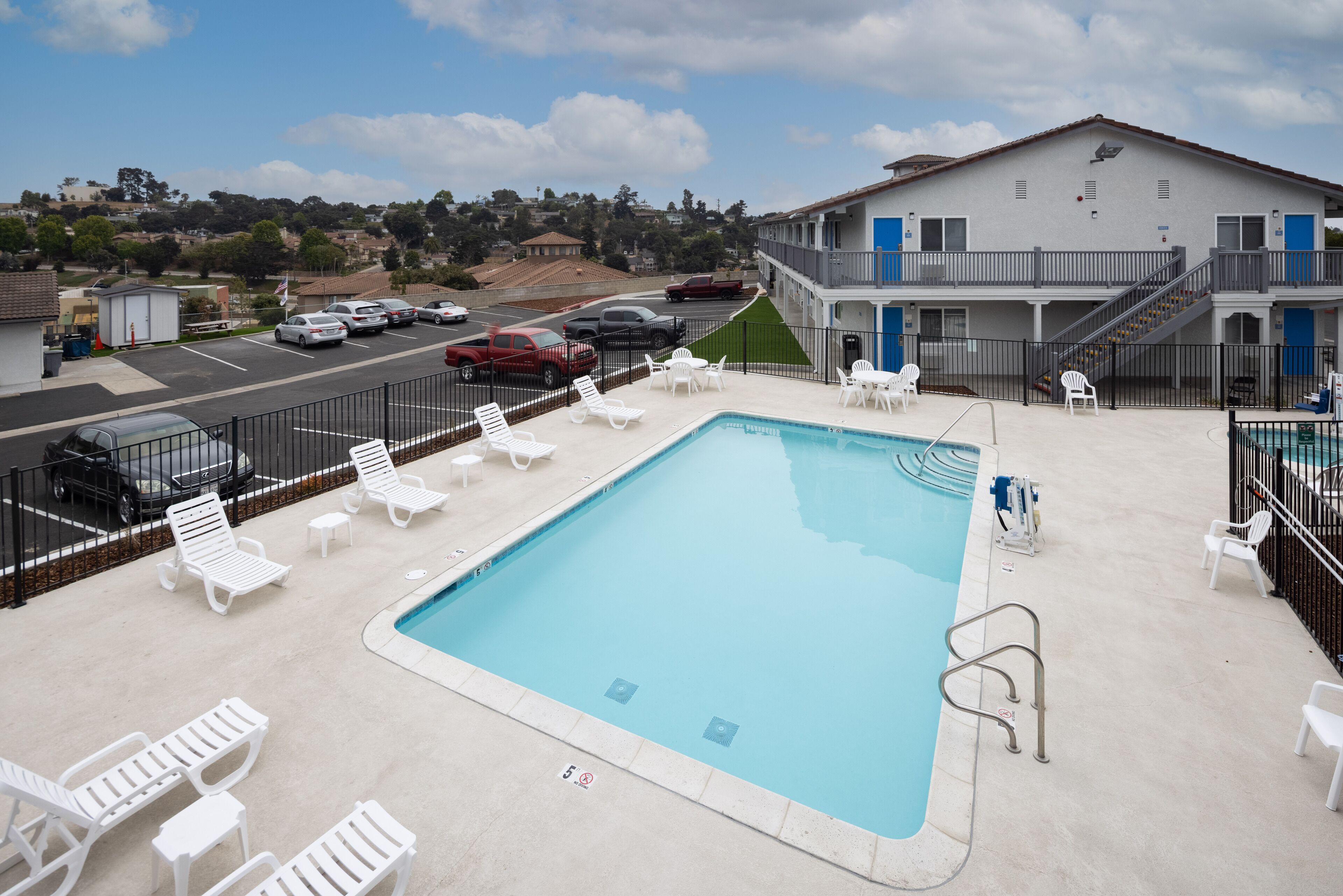 A swimming pool with lounge chairs, a parking lot, and a building with a balcony.