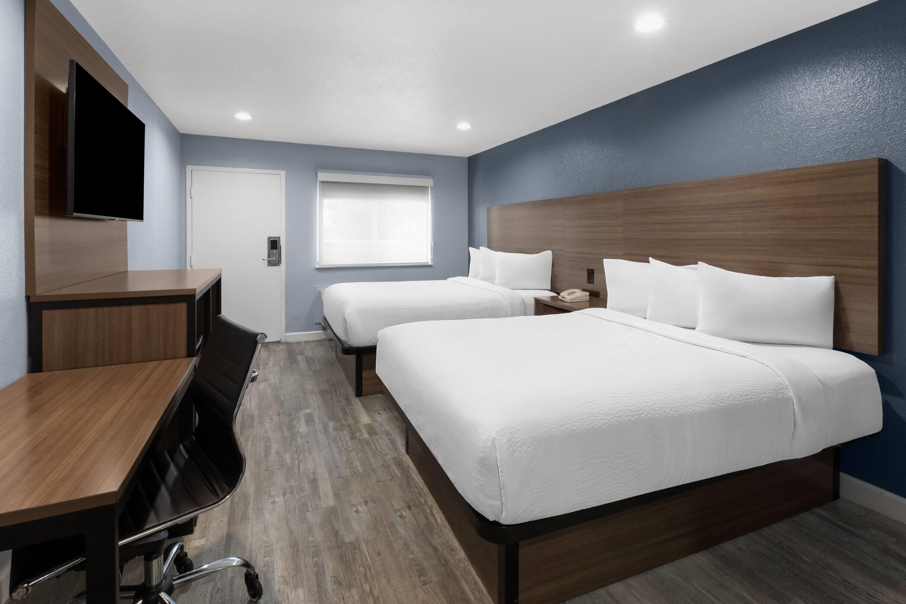 A hotel room with two beds, a desk, a flat-screen TV, and a window with blinds.