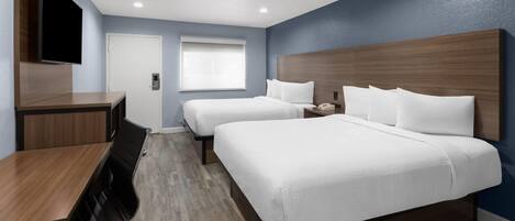 A hotel room with two beds, a desk, a flat-screen TV, and a window with blinds.