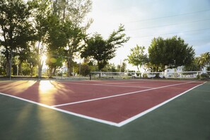An outdoor tennis court with a red surface, surrounded by trees and a white fence. - Topsider Resort (Islamorada)