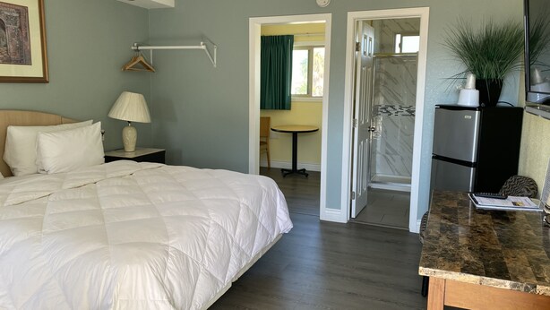 Ein Hotelzimmer mit Bett, Nachttischlampe, einem kleinen Tisch, einem Schreibtisch, einem Kühlschrank und einer Dusche.