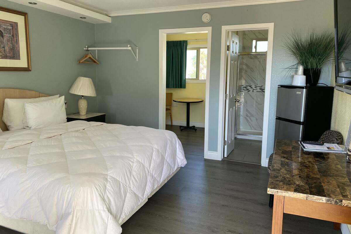 A hotel room with a bed, bedside lamp, a small table, a desk, a refrigerator, and a shower area.