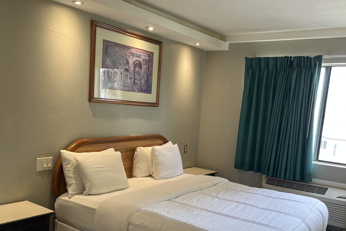 A neatly arranged hotel room with a large bed, a bedside table, a wall-mounted picture, and a window with curtains.