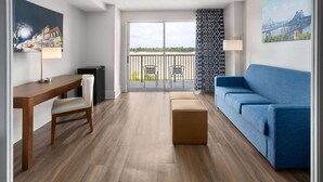 Una sala de estar moderna con piso de madera, un sofá azul, un pequeño reposapiés, un escritorio con una silla, y un balcón con vista a la ciudad.