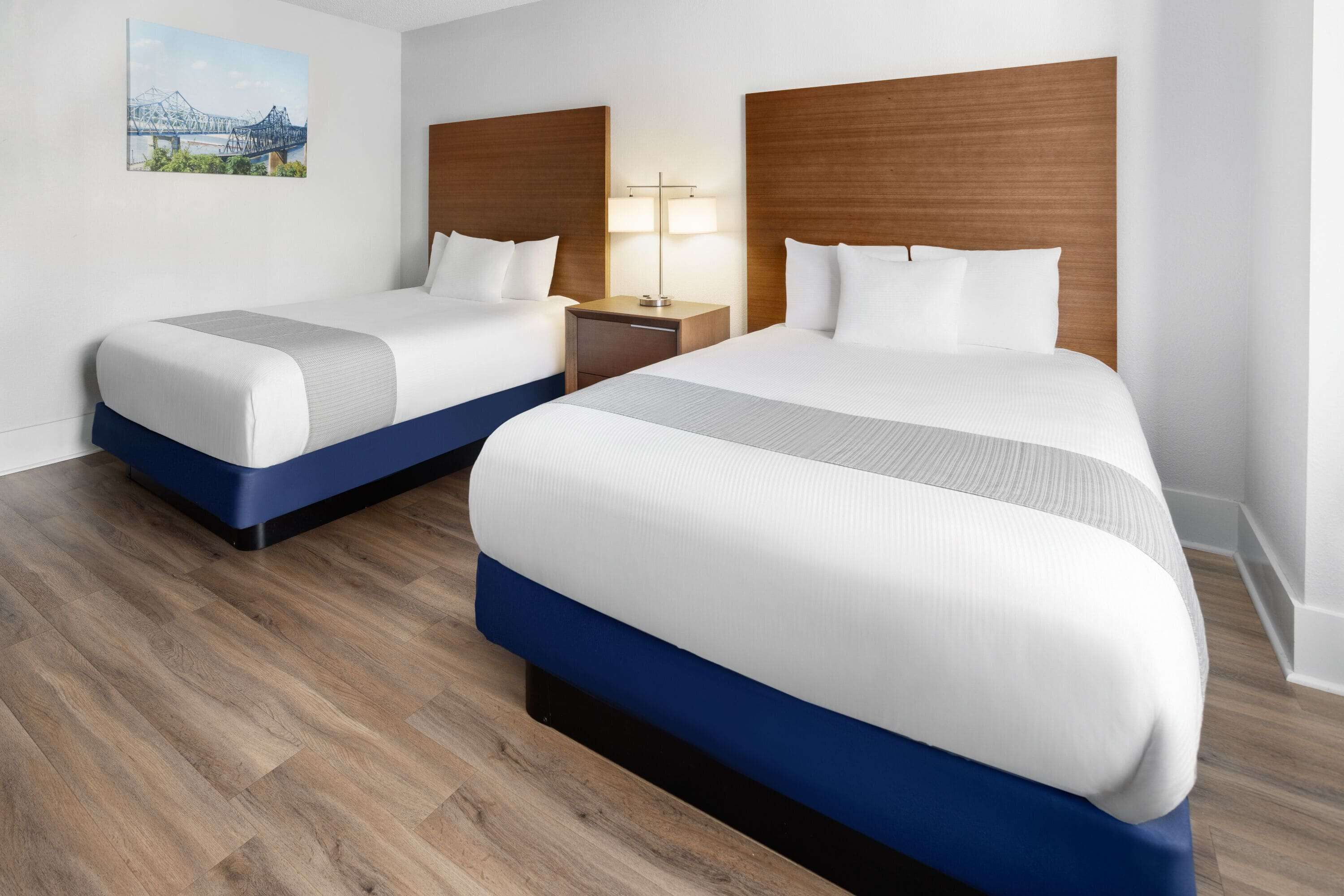 A hotel room with two beds, wooden headboards, a nightstand with a lamp, and a painting on the wall.