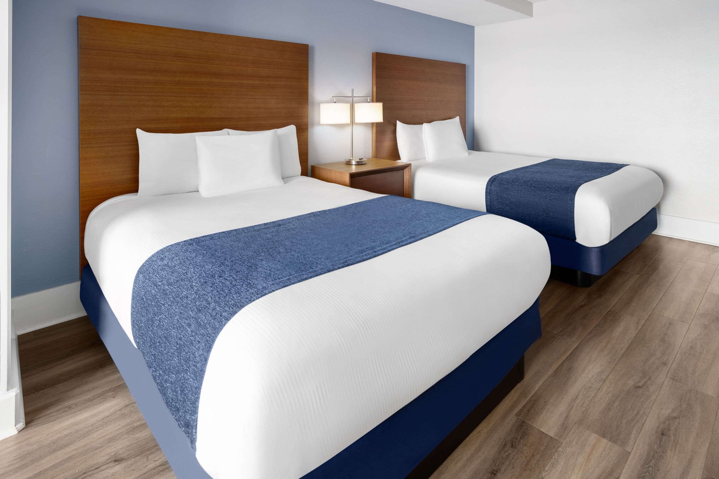 Ein Hotelzimmer mit zwei Betten, Holz-Kopfteilen, einem Nachttisch mit Lampe und einem blauen Akzent an den Betten.