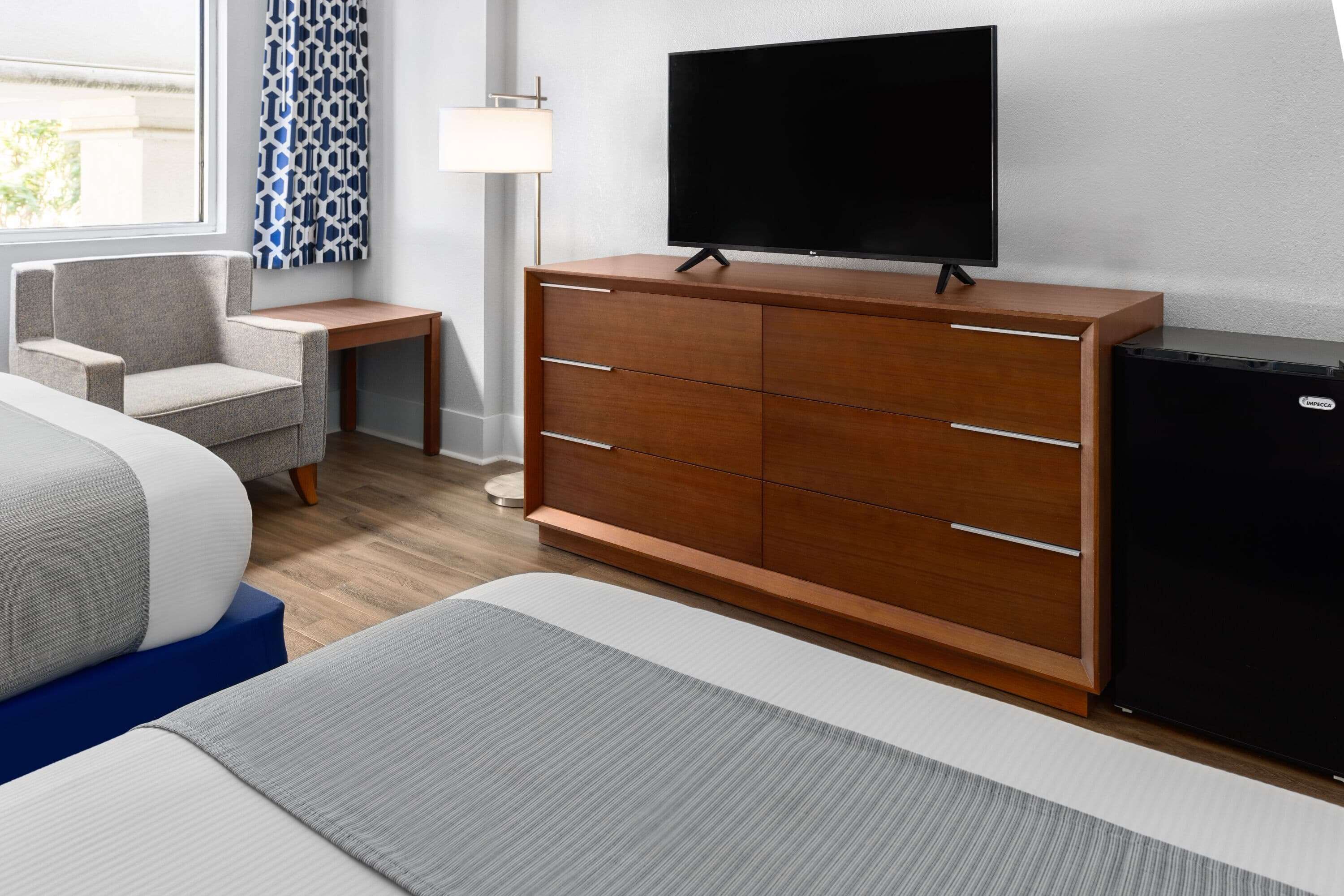 A hotel room with a bed, a wooden dresser, a television, a desk, and a chair.