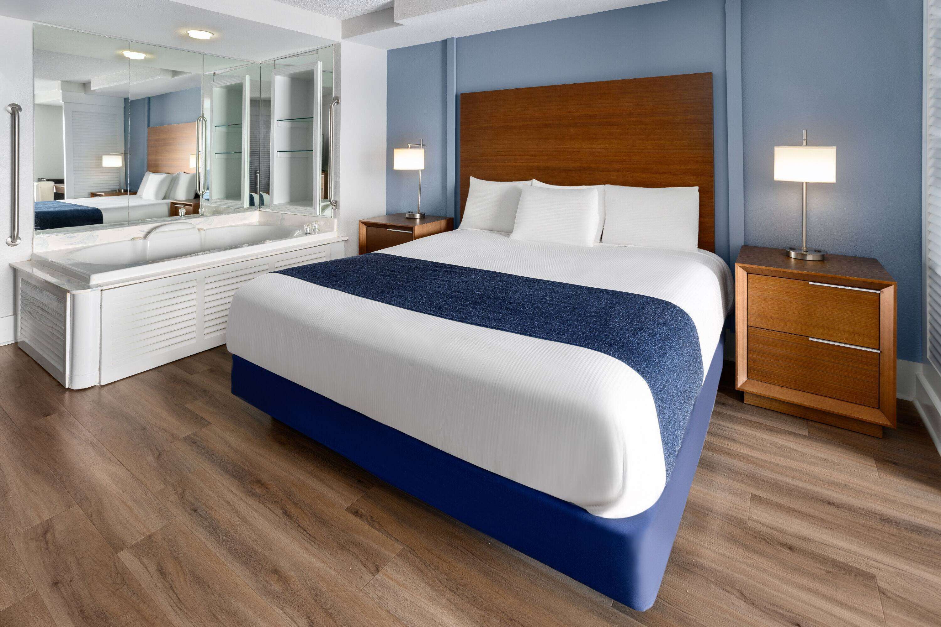 Ein modernes Hotelzimmer mit einem großen Bett, einem separaten Badebereich, einem Holz-Kopfteil, Nachttischlampen und einem großen Spiegel.