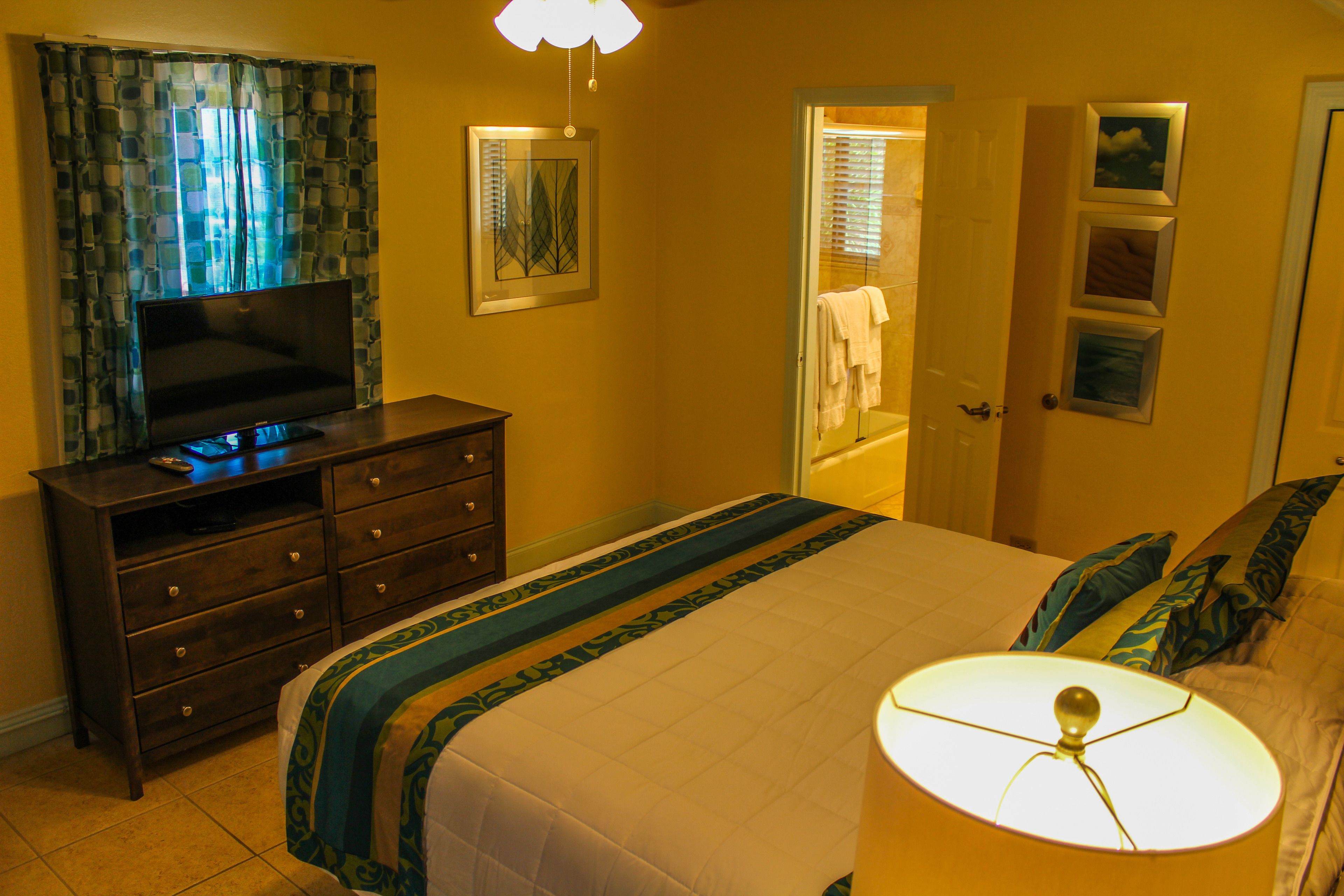 Quarto com cama, cômoda, televisão e banheiro visível por uma porta aberta.