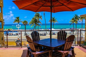 A wooden table and chairs under an umbrella, overlooking a beach with palm trees and the ocean.