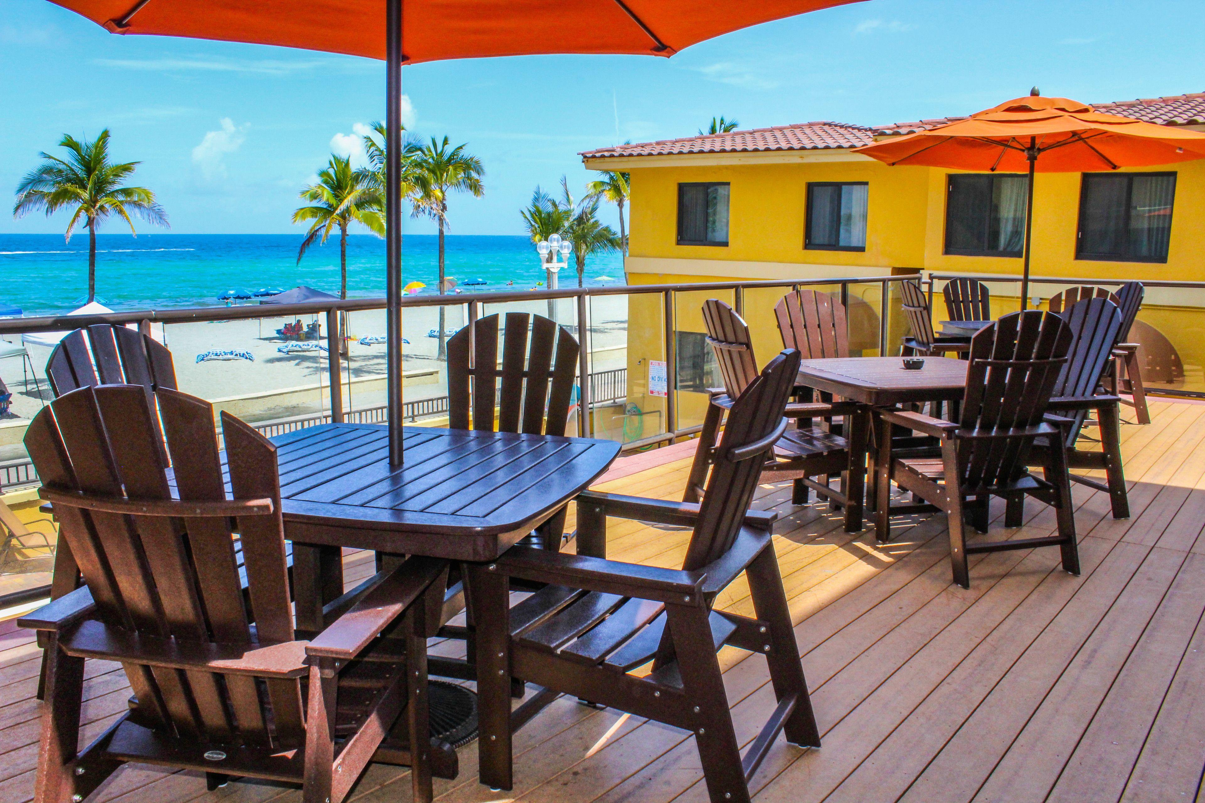 A wooden deck with a table and chairs, overlooking a beach with palm trees and a yellow building.