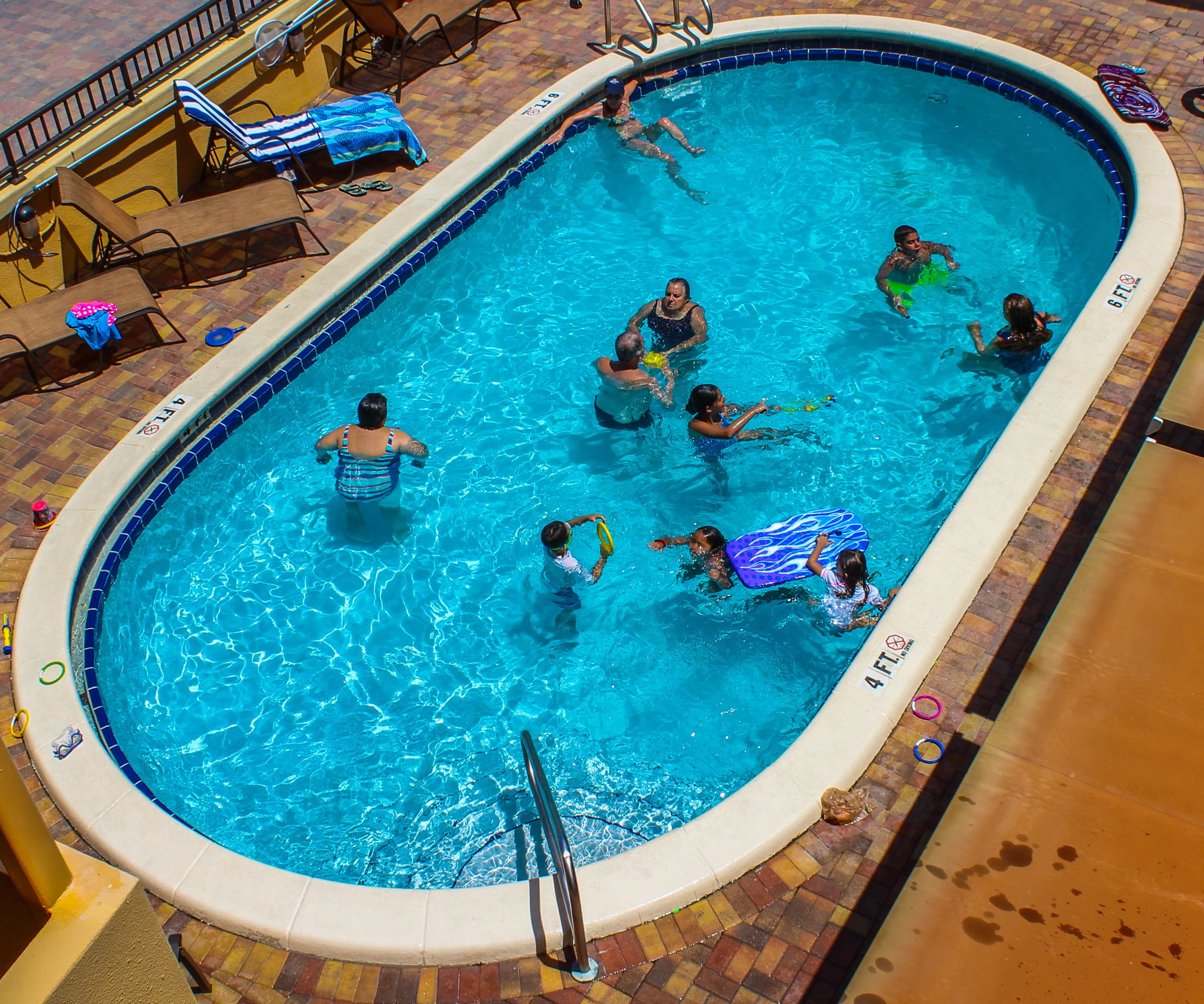 A swimming pool with people in it, surrounded by lounge chairs and a tiled deck.