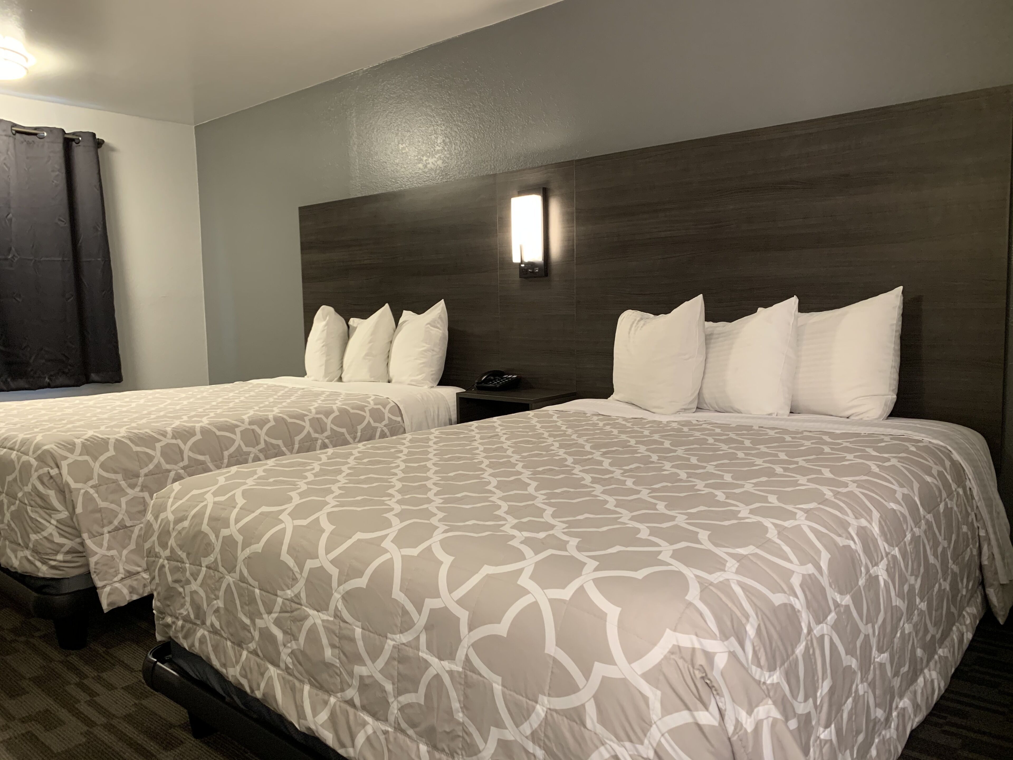 A hotel room with two beds, each with a patterned bedspread and white pillows. There is a nightstand with a phone and a wall-mounted light.