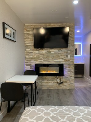 Una sala de estar moderna con televisor de pantalla plana sobre una chimenea de piedra, un comedor con mesa y sillas, y una cocina con encimera y armarios.