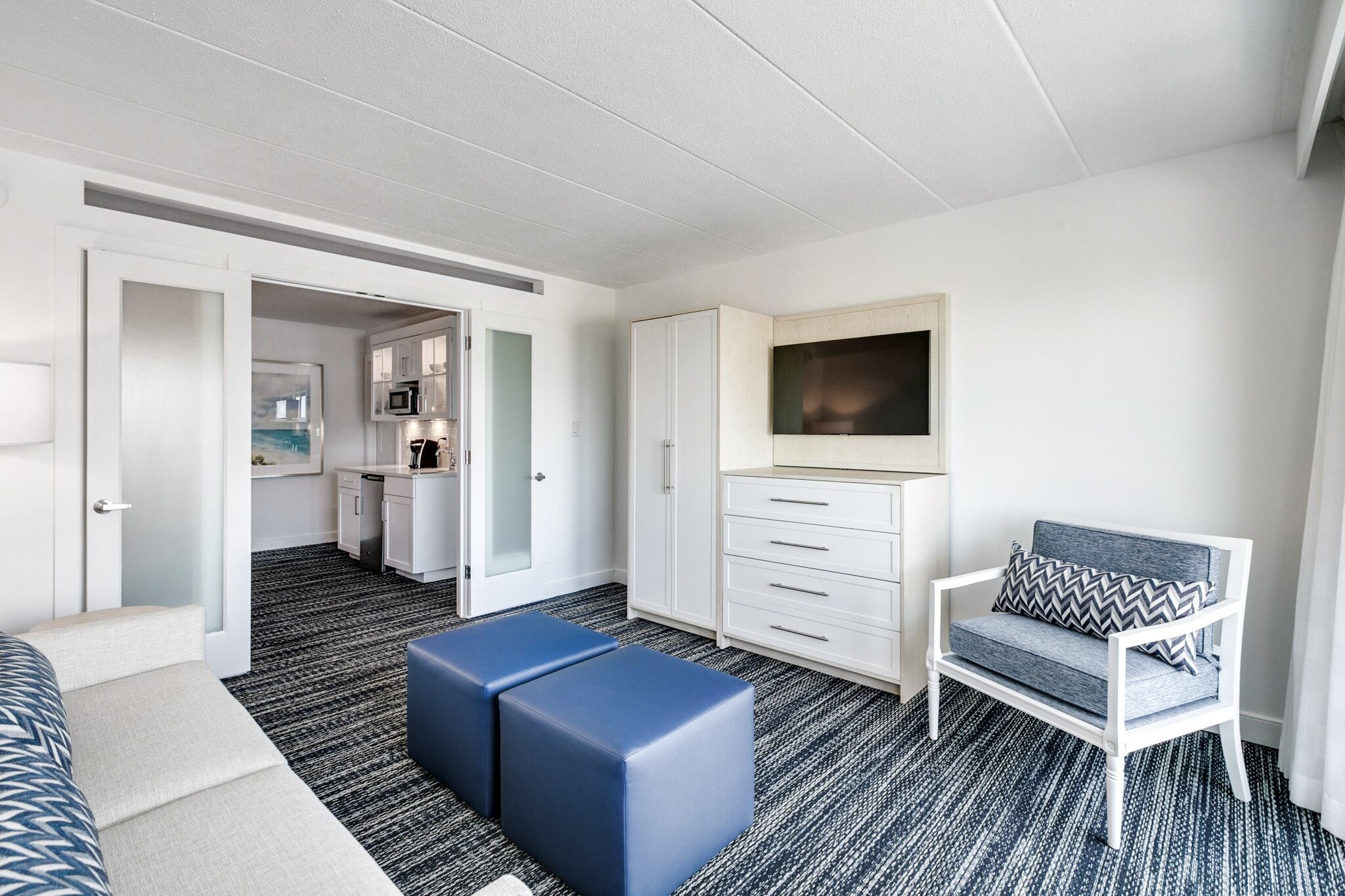 A modern hotel room with a kitchenette, a seating area with a sofa and ottoman, and a TV mounted on the wall.