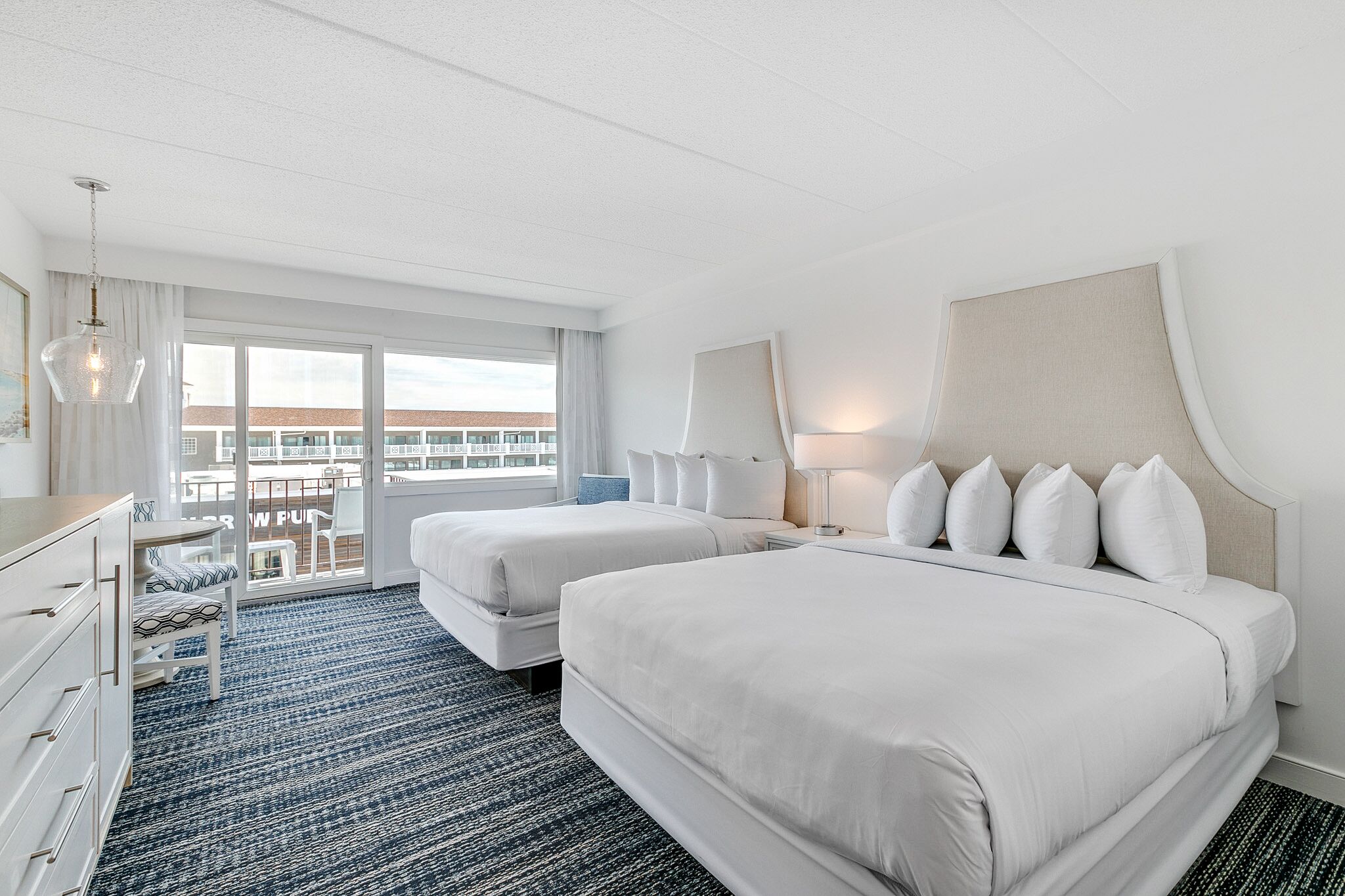 Habitación de hotel con dos camas, un aparador, un escritorio y un ventanal con vistas a edificios.